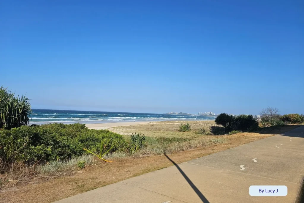Paved beach access path at Bilinga Beach, Gold Coast, with ocean views, coastal shrubs, and distant skyline visible under clear blue skies.