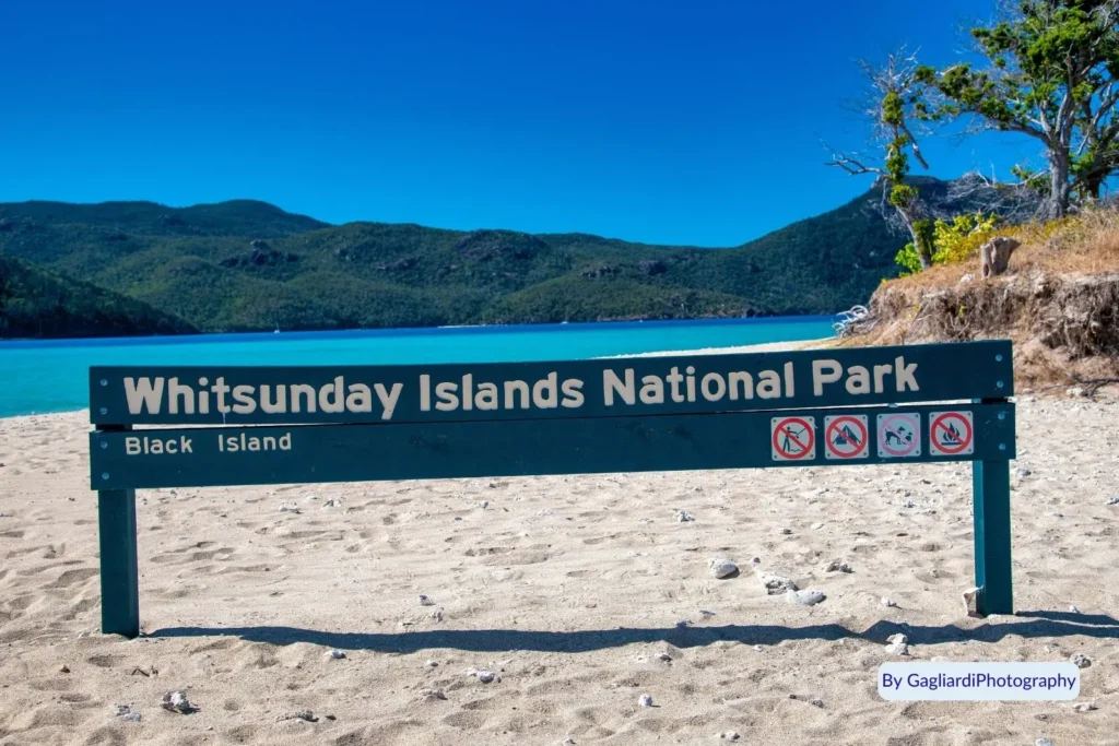 Whitsunday Islands National Park sign on sandy beach at Black Island with turquoise water and green hills in the distance, Queensland, Australia.