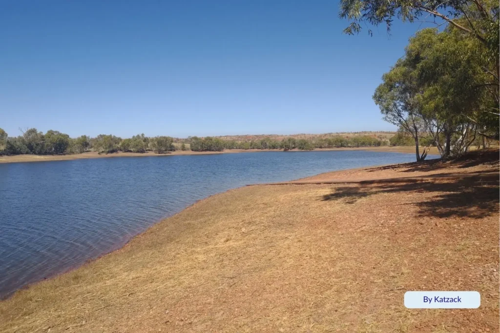 Tranquil riverside scene at Boyne Island with calm blue water, dry grassy shoreline, and scattered gum trees under a clear blue Queensland sky.