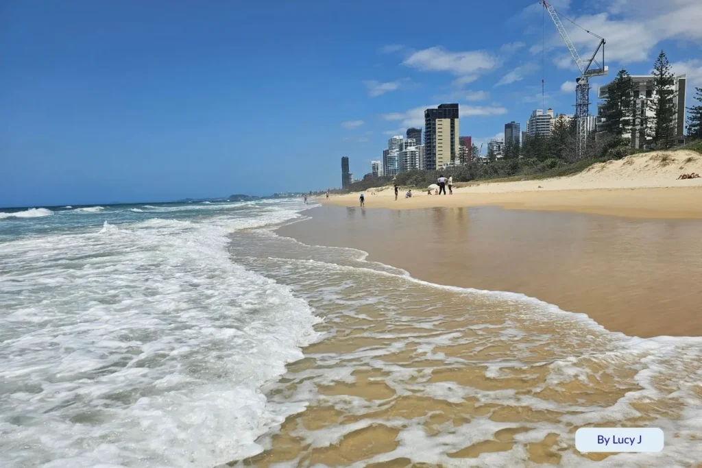 Foamy waves washing onto golden sand at Broadbeach, Gold Coast, with people walking along the shoreline and high-rise buildings in the background.