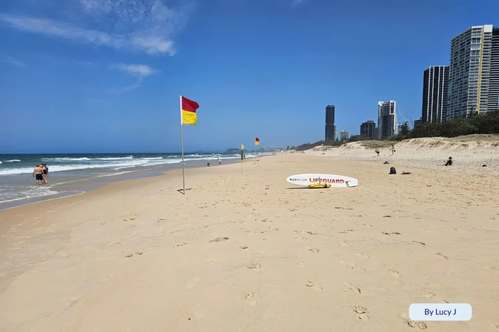 Patrolled swimming area at Broadbeach, Gold Coast, with red and yellow lifeguard flags, soft sand, and tall beachfront towers under a sunny blue sky.