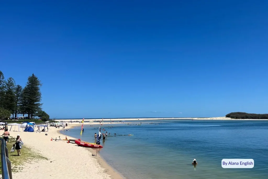 Families swimming and relaxing on the sandy foreshore of Bulcock Beach, Caloundra, with calm turquoise water and views across Pumicestone Passage on the Sunshine Coast, Queensland.
