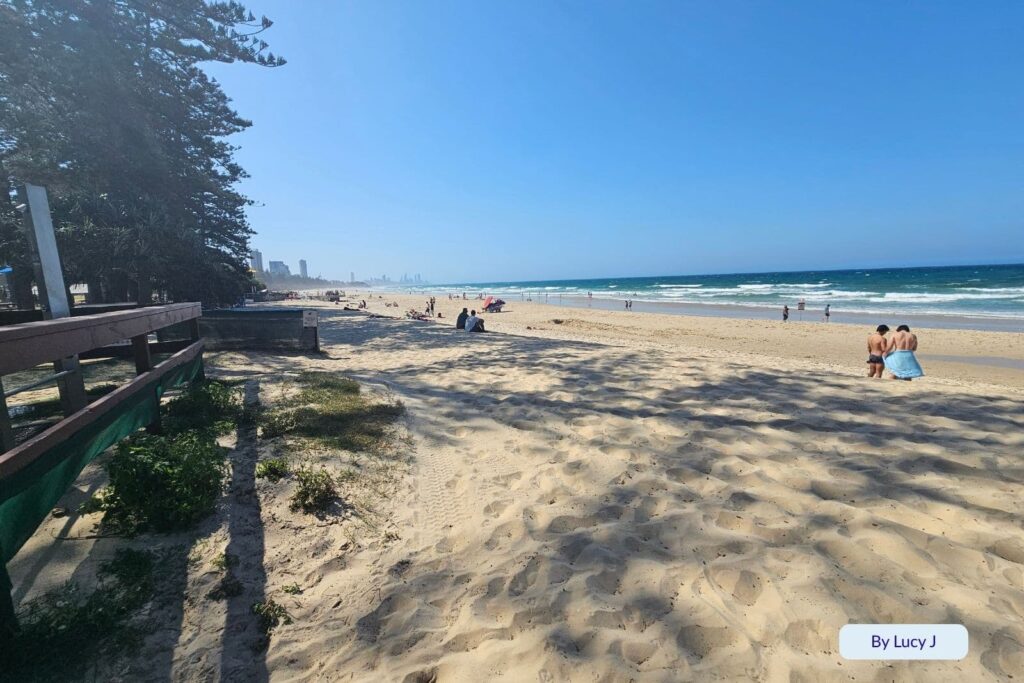 Wide sandy stretch at Burleigh Heads Beach, Gold Coast, with scattered beachgoers, distant skyline views, and shaded foreshore seating.