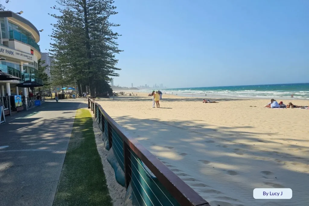 Pathway beside Burleigh Heads Beach with ocean views, beachfront shops, and people walking along the sand under tall Norfolk pines.