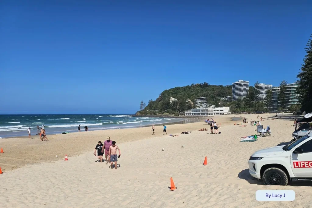 Families enjoying a sunny day at Burleigh Heads Beach, Gold Coast, with surf patrol cones, soft sand, and Burleigh Headland rising in the background.