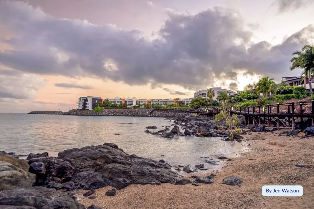 Rocky foreshore and calm ocean at Cannonvale Beach during sunset with colourful sky and coastal apartments, Whitsundays, Queensland.