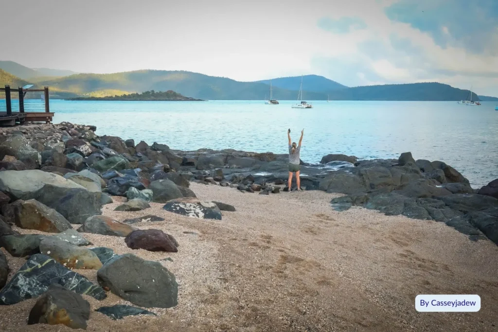 Woman standing on rocky shore overlooking turquoise bay and moored boats at Cannonvale Beach, near Airlie Beach, Whitsundays, Queensland.