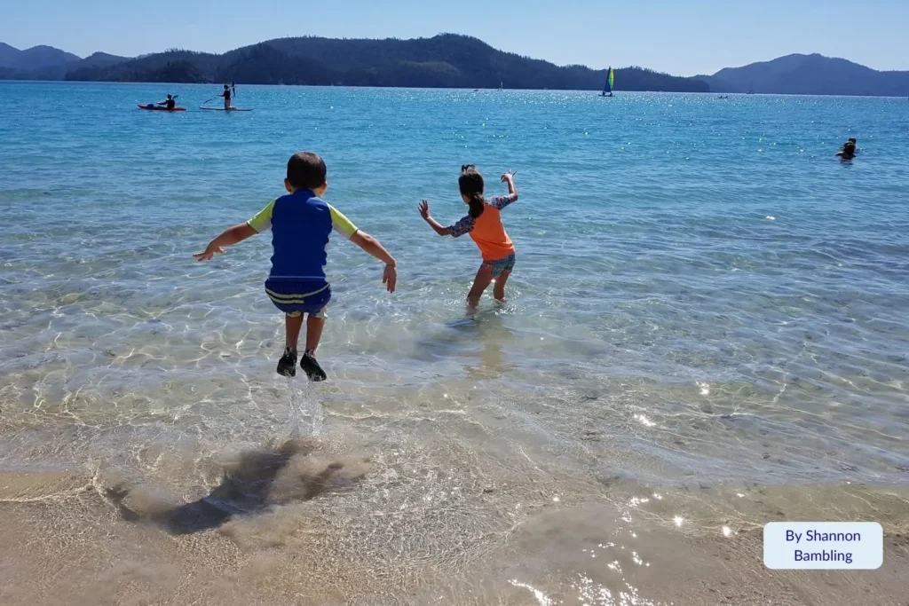 Children playing in the shallow turquoise water at Catseye Beach with sailboats and Whitsunday Islands in the distance, Hamilton Island, Queensland.