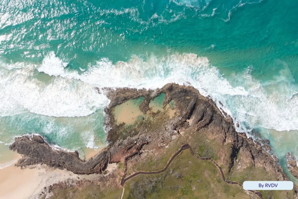 Aerial view of Champagne Pools, Fraser Island (K’gari), showing waves breaking over rock ledges and crystal-blue water along the coastline