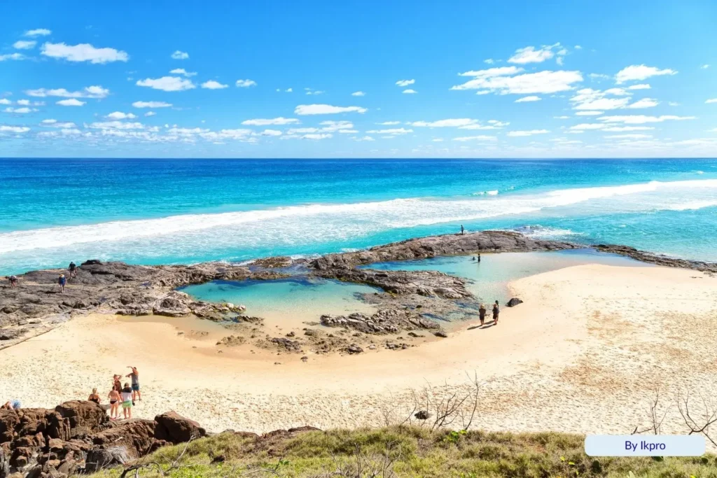 Visitors relaxing at Champagne Pools on Fraser Island (K’gari), where ocean waves spill over volcanic rocks into calm turquoise pools beneath a blue sky