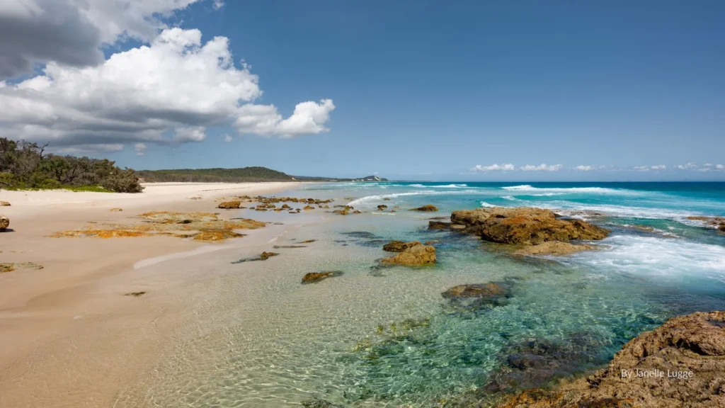 Shallow rock pools at Champagne Pools, Moreton Island with golden sand, clear water, and scattered seaweed under a cloudy blue sky.