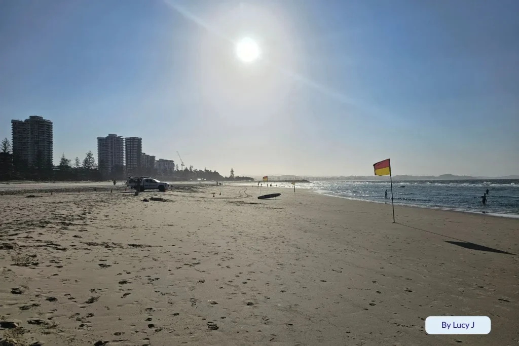 Sunset view over Coolangatta Beach with a lifeguard flag, footprints in the sand, and high-rise buildings silhouetted in the golden light.