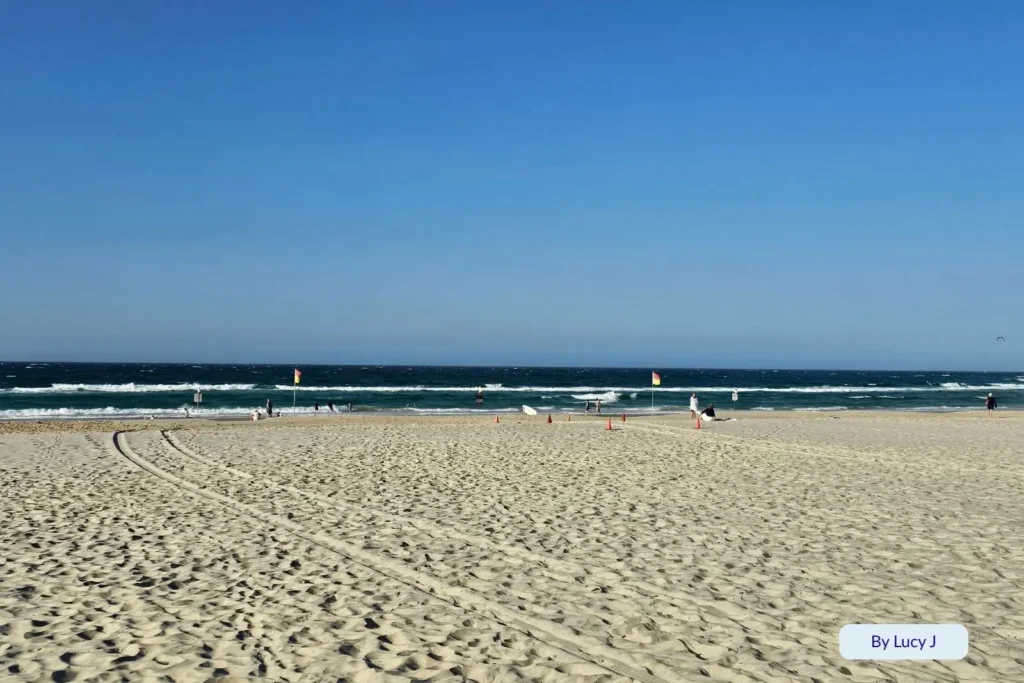 Wide sandy shoreline at Coolangatta Beach with tire tracks in the foreground, surf patrol flags in the distance, and gentle waves under a clear blue sky.