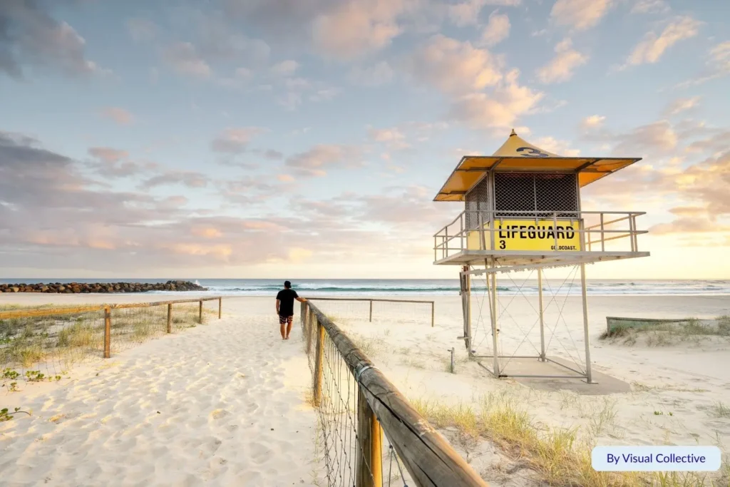 Man walking toward the surf past the yellow lifeguard tower at sunrise on Coolangatta Beach, Gold Coast, Queensland, with golden light and calm waves.