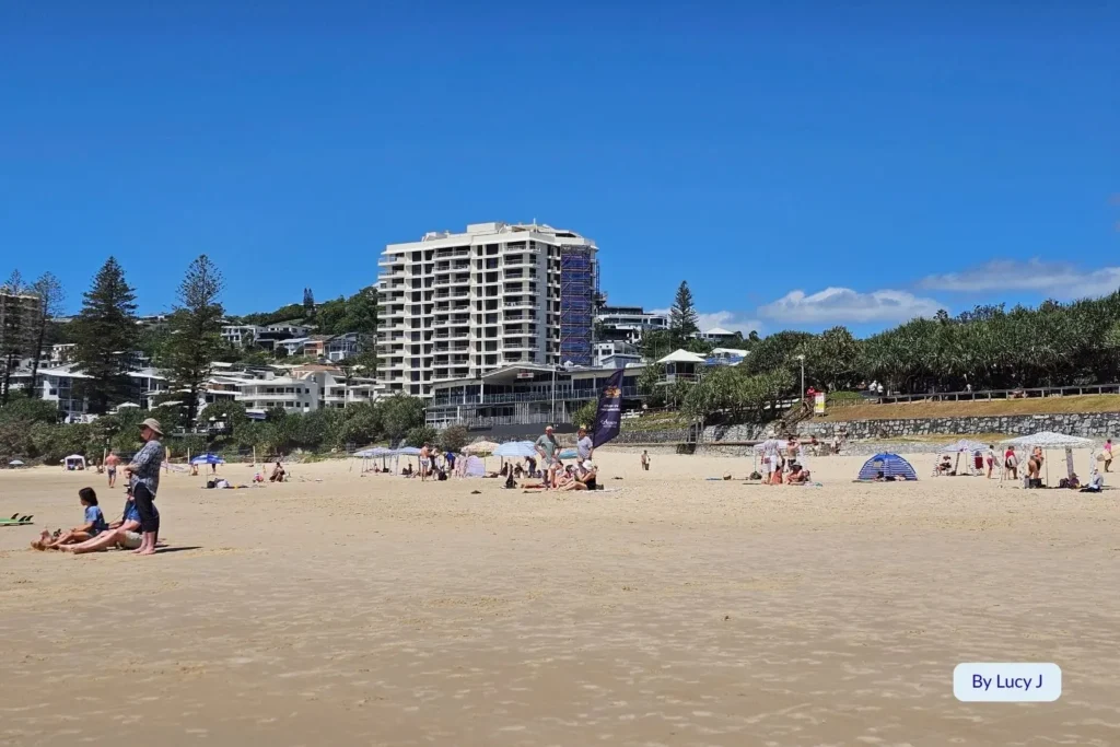 Visitors relaxing on the wide golden sands of Coolum Beach, Sunshine Coast, Queensland, with oceanfront apartments and pandanus trees under a clear blue sky.