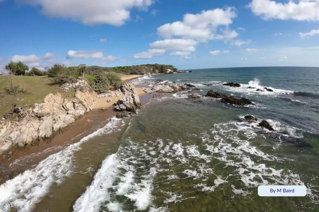 Rocky coastline and small sandy cove at Joey Lee’s Rest Beach on Curtis Island, near Gladstone, Queensland, with clear blue water, foamy waves, and green headlands under a bright sky.
