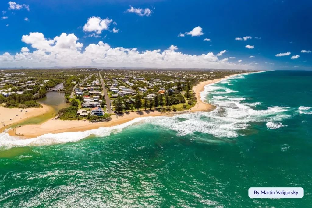 Panoramic aerial of Dicky Beach and Pumicestone Passage, Sunshine Coast, Queensland — golden beach, rolling surf, and suburban coastline under bright blue skies.