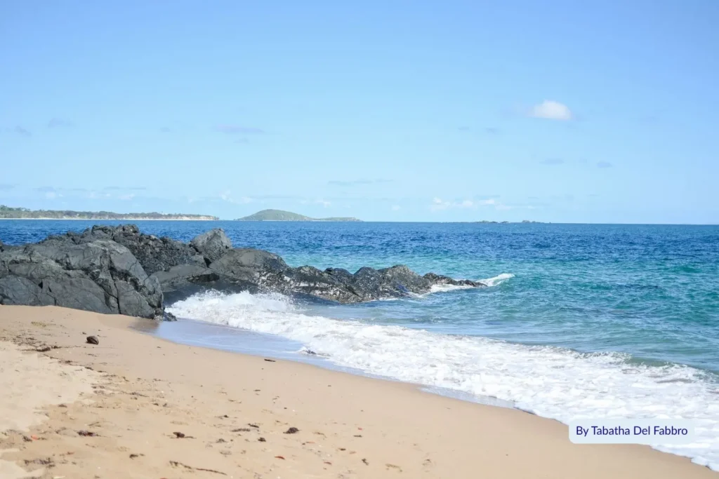 Waves rolling over dark volcanic rocks onto the sandy shore at Eimeo Beach near Mackay, Queensland, under bright blue skies.