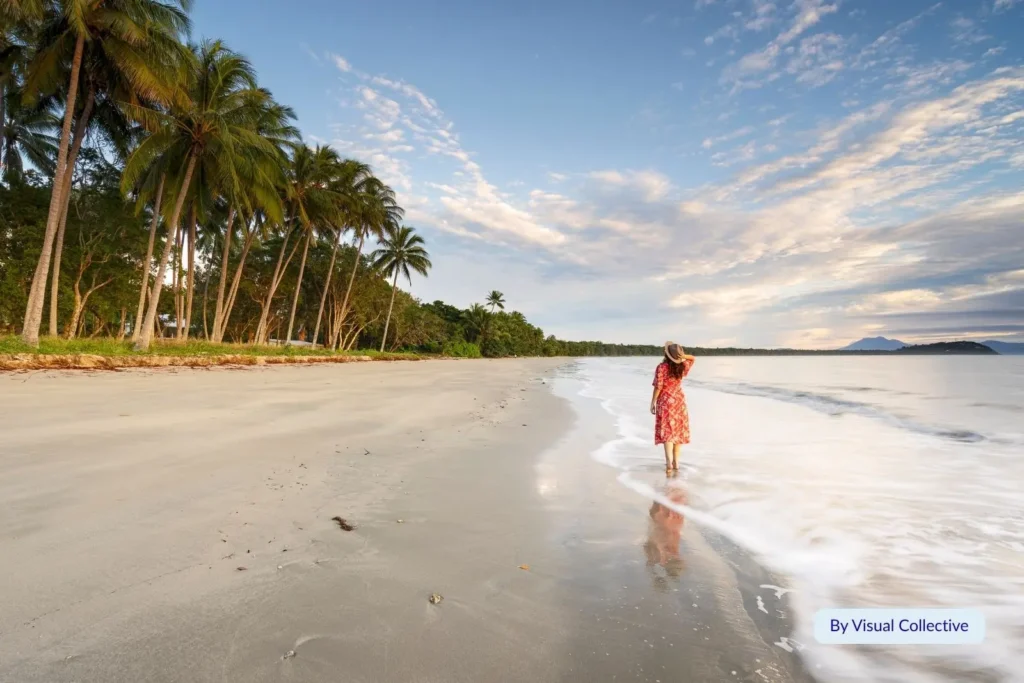 Woman walking along the wet sand of Four Mile Beach at sunrise surrounded by palm trees and coral sea views, Port Douglas, Queensland, Australia