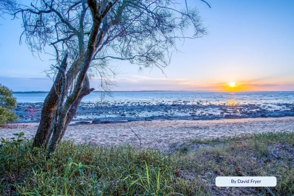 Golden sunset at Gatakers Bay Beach Hervey Bay, with soft evening light over the rocky shoreline and tranquil sea, viewed through coastal trees.