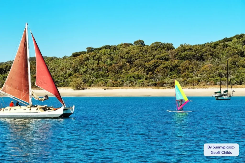 Sailboat with red sails and windsurfer on the blue waters off Great Keppel Island, Queensland, near a forested sandy shore.