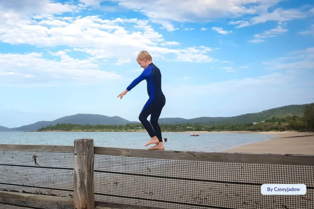 Child balancing on a wooden fence overlooking the calm waters at Harbour Beach in Mackay, Queensland, with green headlands in the distance