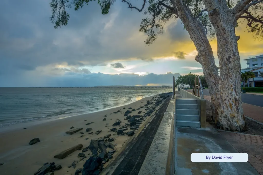Sunset along Hervey Bay Esplanade with calm seas, rock walls, and a large tree framing the coastal pathway.