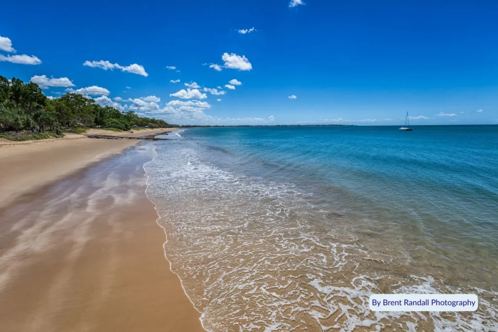 Golden sand shoreline and calm turquoise waters along Hervey Bay Esplanade under a bright blue Queensland sky.