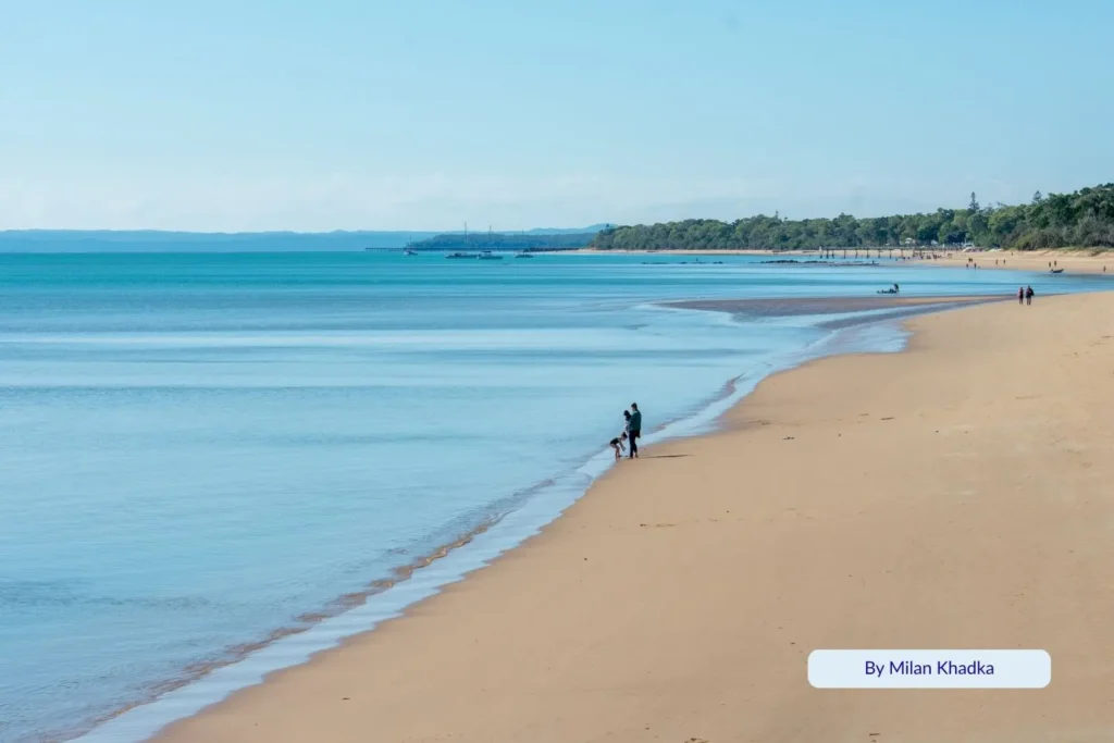 Wide sandy beach at Hervey Bay with a lone fisherman standing near the gentle waves on a sunny day.