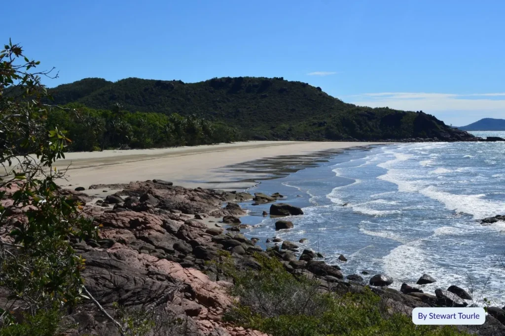 Rocky shoreline and gentle surf at Hinchinbrook Island National Park with lush green headlands and blue sky, Queensland
