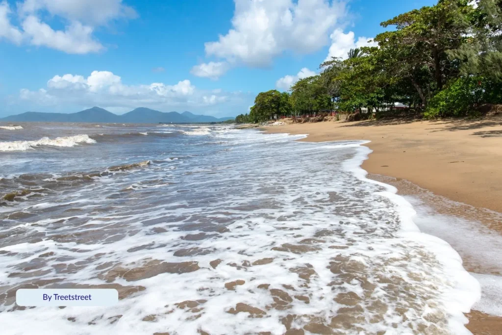 Gentle waves rolling onto the sandy shoreline of Holloways Beach, Cairns, with palm trees and coastal mountains in the background.