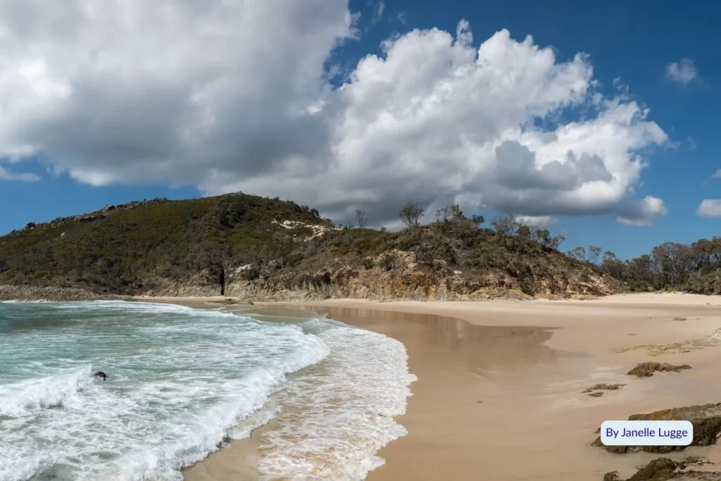 Small secluded beach at Honeymoon Bay Moreton Island showing gentle surf, golden sand, and green hills in the background on a sunny day.