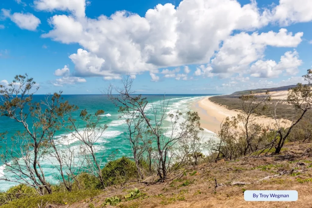 Aerial view of Indian Head on Fraser Island (K’gari) with 4WD vehicles driving along the golden sands of 75 Mile Beach under a clear blue sky.