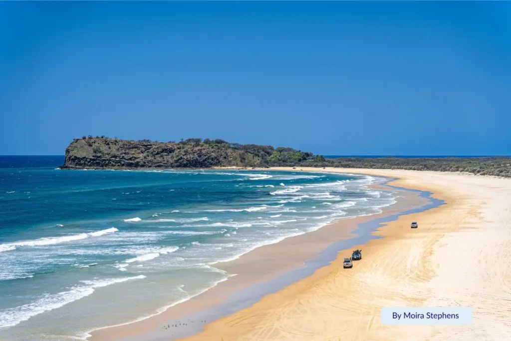 Aerial view of Indian Head on Fraser Island (K’gari) with 4WD vehicles driving along the golden sands of 75 Mile Beach and turquoise surf rolling in from the Coral Sea