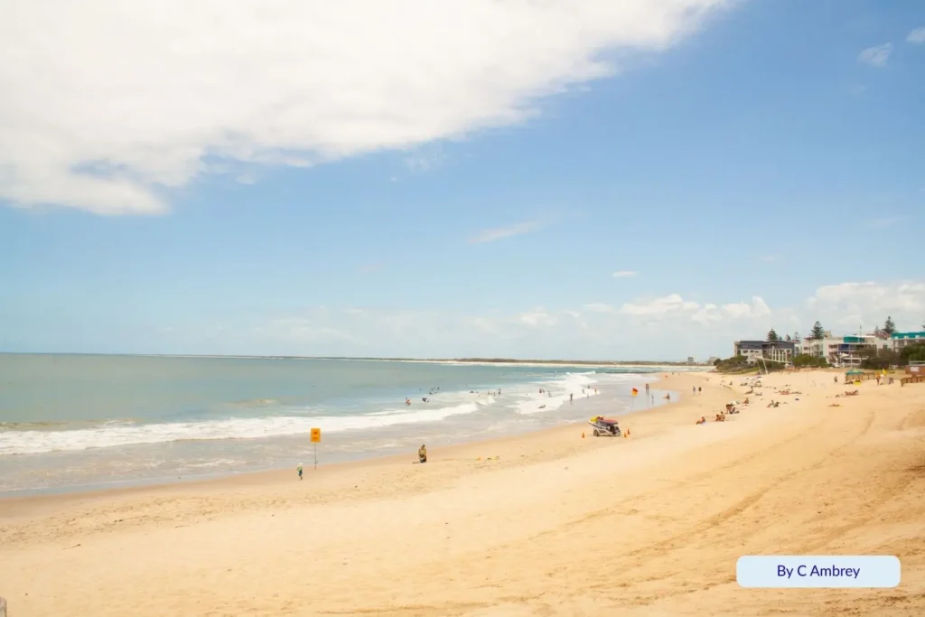 Sunny day at Kings Beach, Caloundra, Queensland — families relaxing on the wide sandy beach with gentle surf and the town skyline in the background.