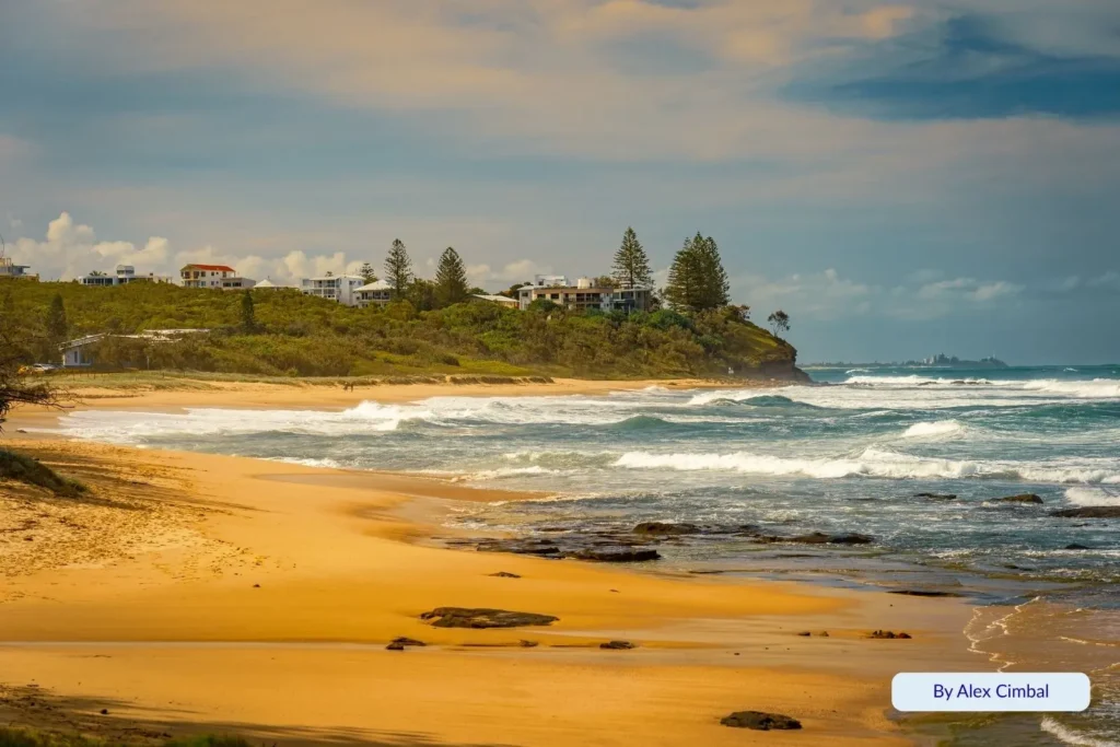 Rocky headland and waves at Kings Beach, Sunshine Coast, Queensland — dramatic shoreline with surf, golden sand, and houses perched on the hill in the distance.
