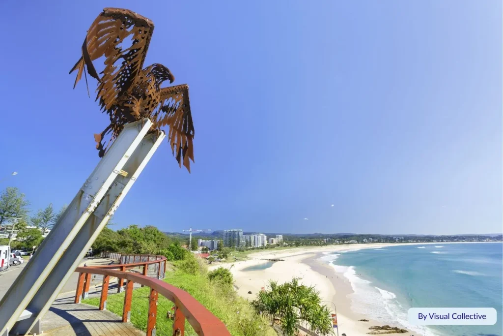 Metal eagle sculpture overlooking the sweeping curve of Kirra Beach with golden sand, surf waves, and the Coolangatta coastline in the distance, Gold Coast, Queensland.