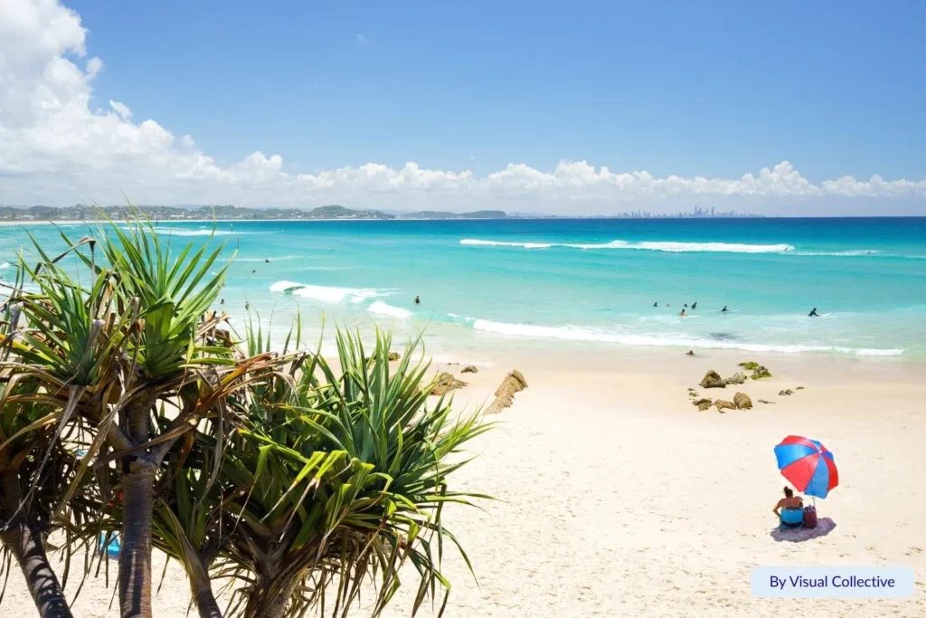 Pandanus trees framing the white sand and turquoise waves of Kirra Beach, Gold Coast, Queensland, with swimmers and a red-and-blue beach umbrella on the shore.
