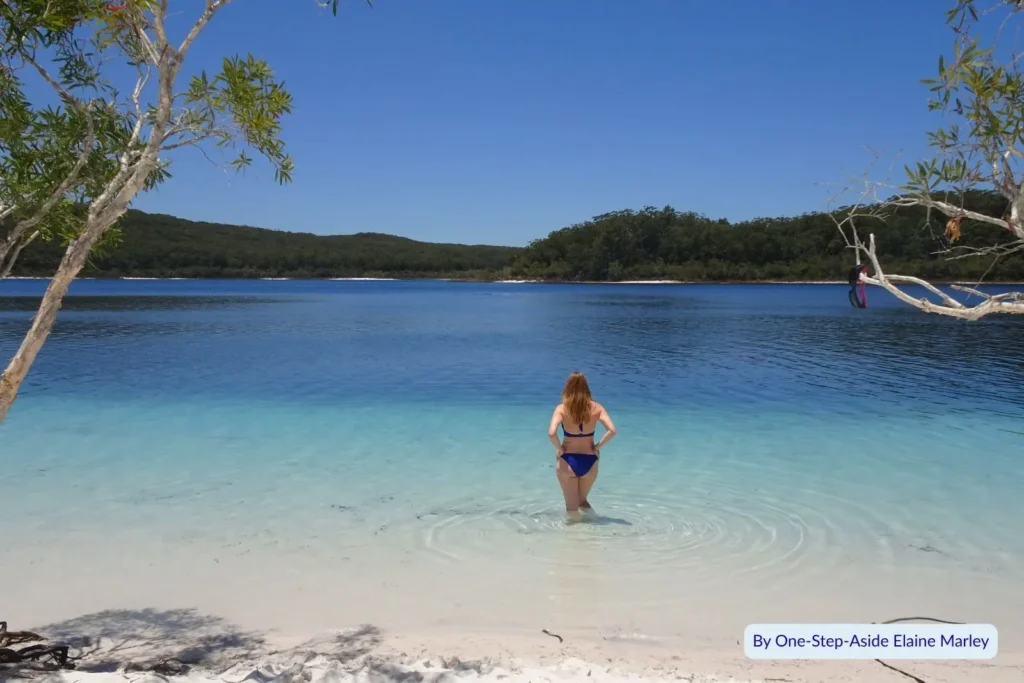 Visitor standing in the calm blue shallows of Lake McKenzie with white sandy beach and forest backdrop on Fraser Island (K’gari), Queensland.