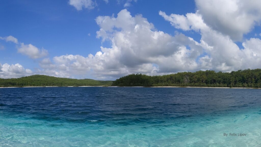Deep blue freshwater of Lake McKenzie under fluffy white clouds, framed by green forest hills on Fraser Island (K’gari), Queensland.