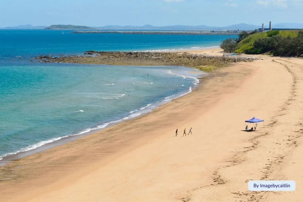 Aerial view of Lamberts Beach, Mackay, Queensland, showing clear turquoise water, gentle waves, and a small group relaxing under a beach umbrella