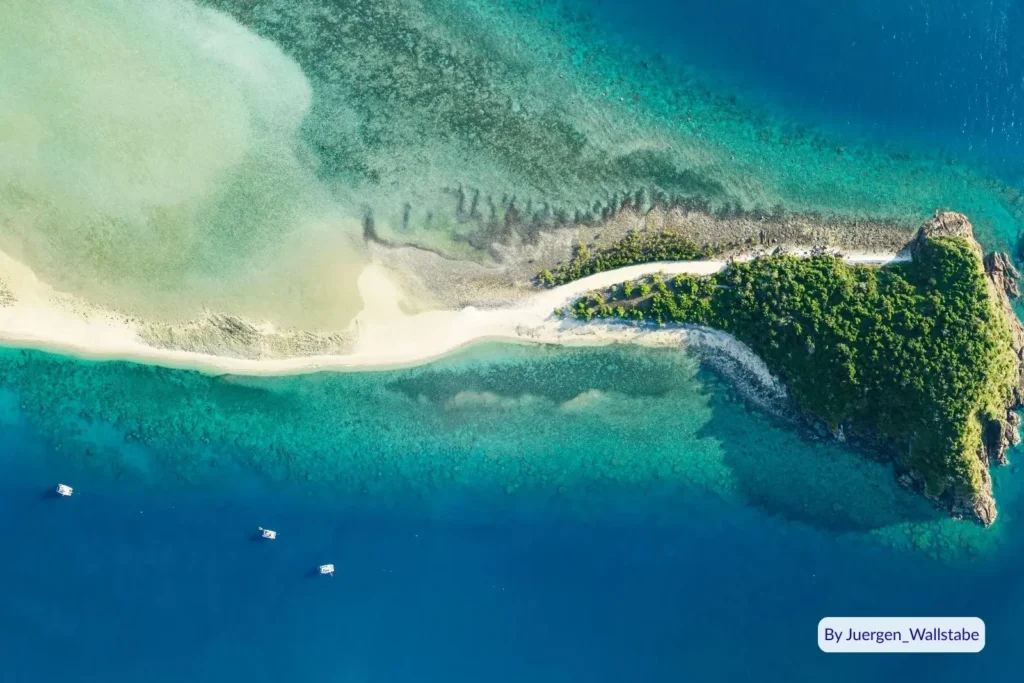 Drone view of Langford Island’s narrow sand spit and surrounding coral reef lagoon with turquoise waters and anchored boats, Whitsundays, Great Barrier Reef, Queensland, Australia.
