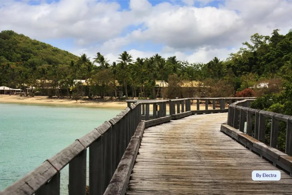 Wooden boardwalk curving along turquoise water toward palm-fringed Palm Bay Resort on Long Island, Whitsundays, Queensland, Australia