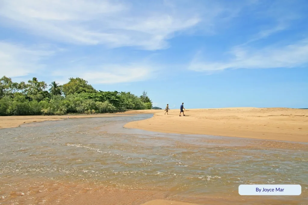 Two people walking along the sandy shoreline at Machans Beach, Cairns, Queensland, where a tidal creek meets the ocean under a bright blue sky with lush tropical vegetation in the background.