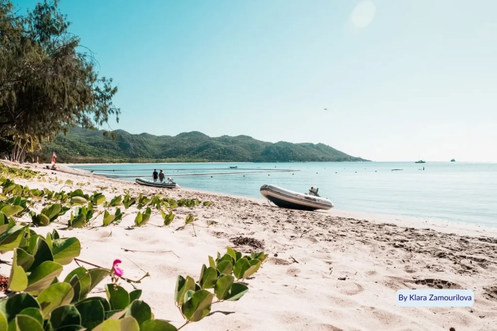 Sunny day on Magnetic Island with white sandy beach, small boats on shore, and mountain backdrop, North Queensland.