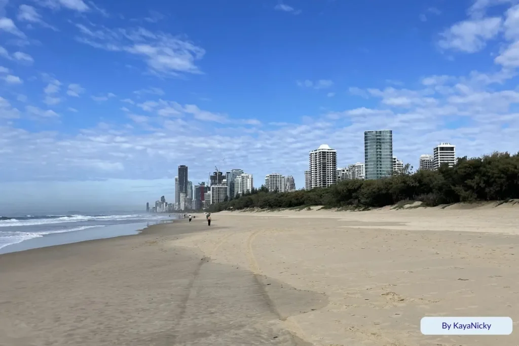 Wide sandy shoreline at Main Beach with gentle surf, coastal dunes, and the Gold Coast skyline in the distance under a bright blue sky, Queensland, Australia.
