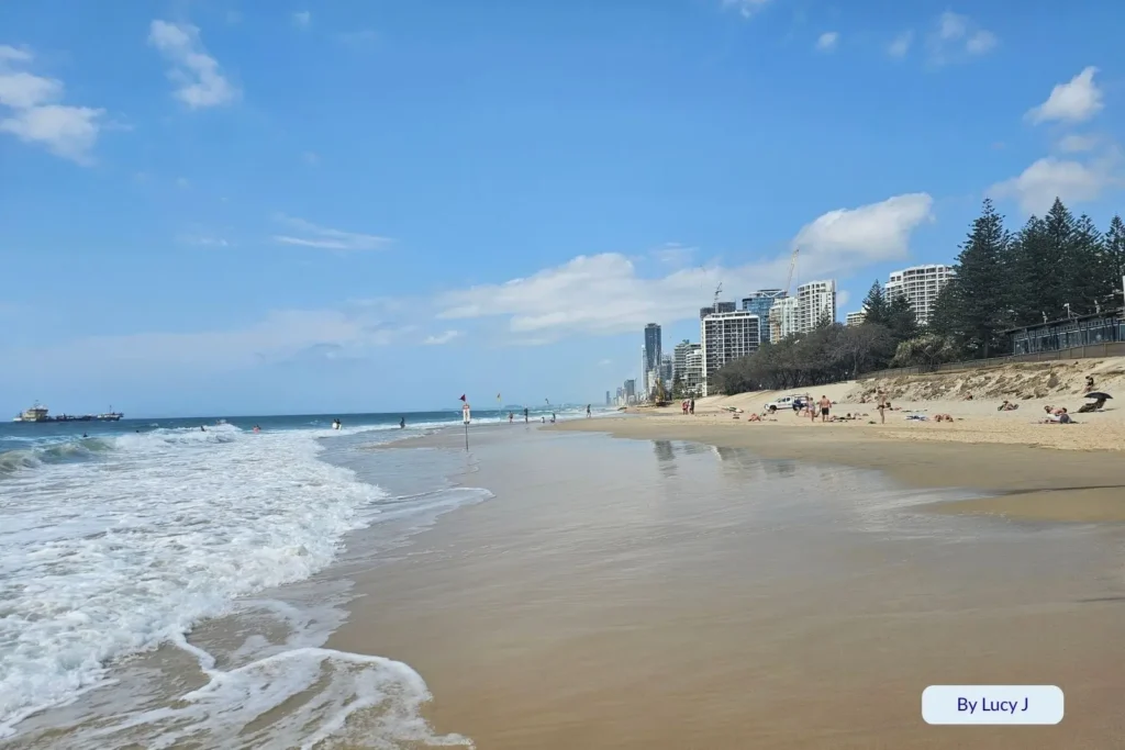 View along Main Beach, Southport, showing surf, shoreline reflections, and nearby Gold Coast skyline under bright blue skies, Queensland, Australia
