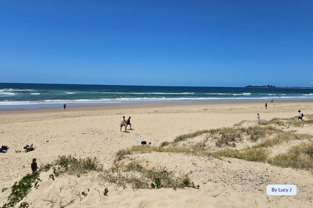 People relaxing and walking along the golden sand at Maroochydore Beach, Sunshine Coast, Queensland, with clear blue water and grassy dunes in the foreground.