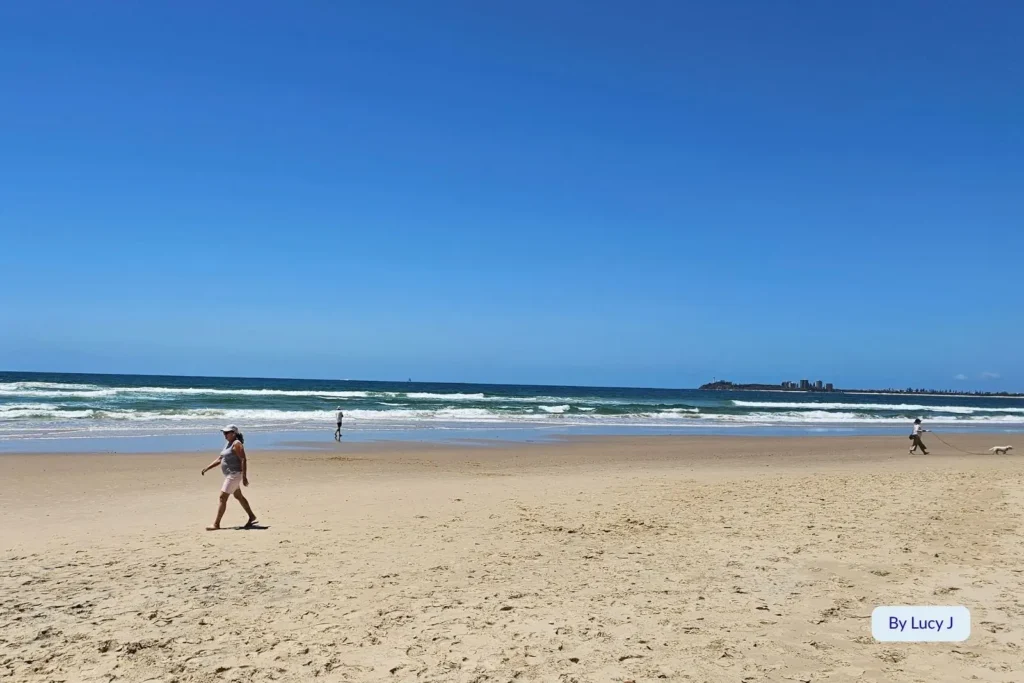 Wide sandy shoreline and gentle surf at Maroochydore Beach, Sunshine Coast, Queensland, with beach walkers enjoying a sunny day and Mudjimba Island in the distance.