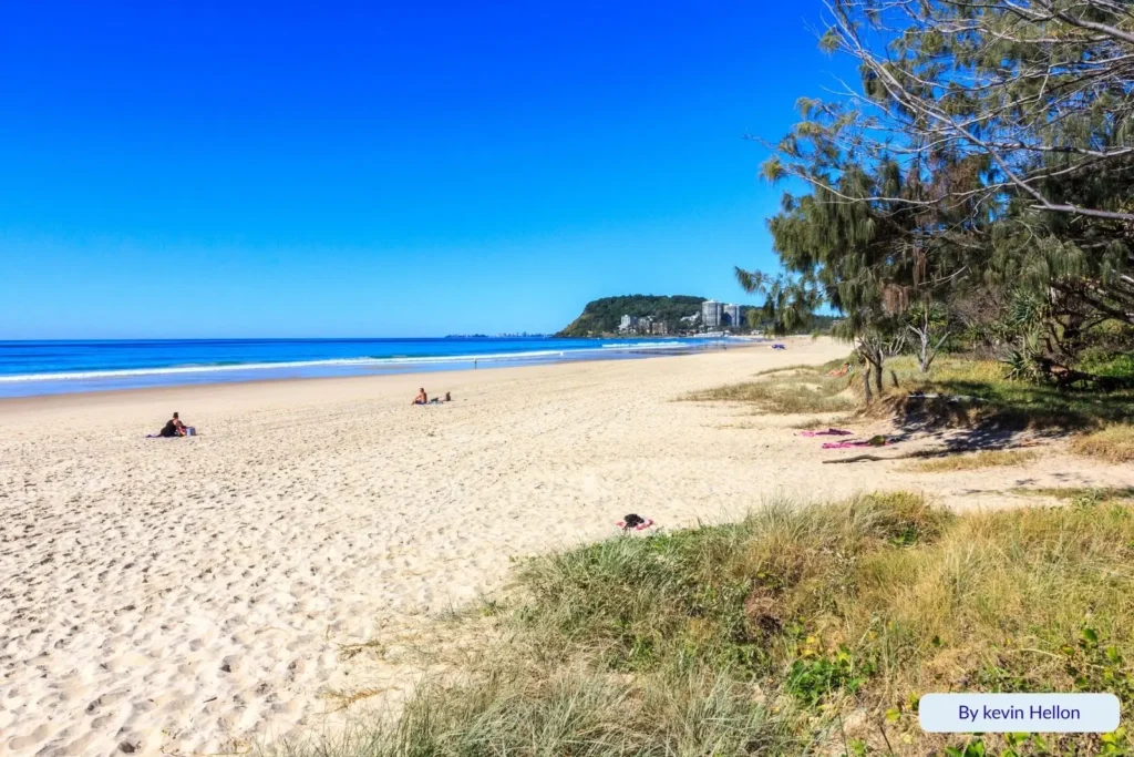 Wide stretch of Mermaid Beach with golden sand, gentle surf, and Burleigh Headland visible in the distance under a bright blue sky, Gold Coast, Queensland.